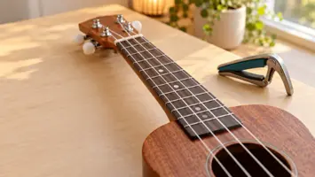 Gripebrettet på en ukulele sett fra spillerens vinkel, med fire strenger og posisjonsprikker, og en capo på bordet ved siden av